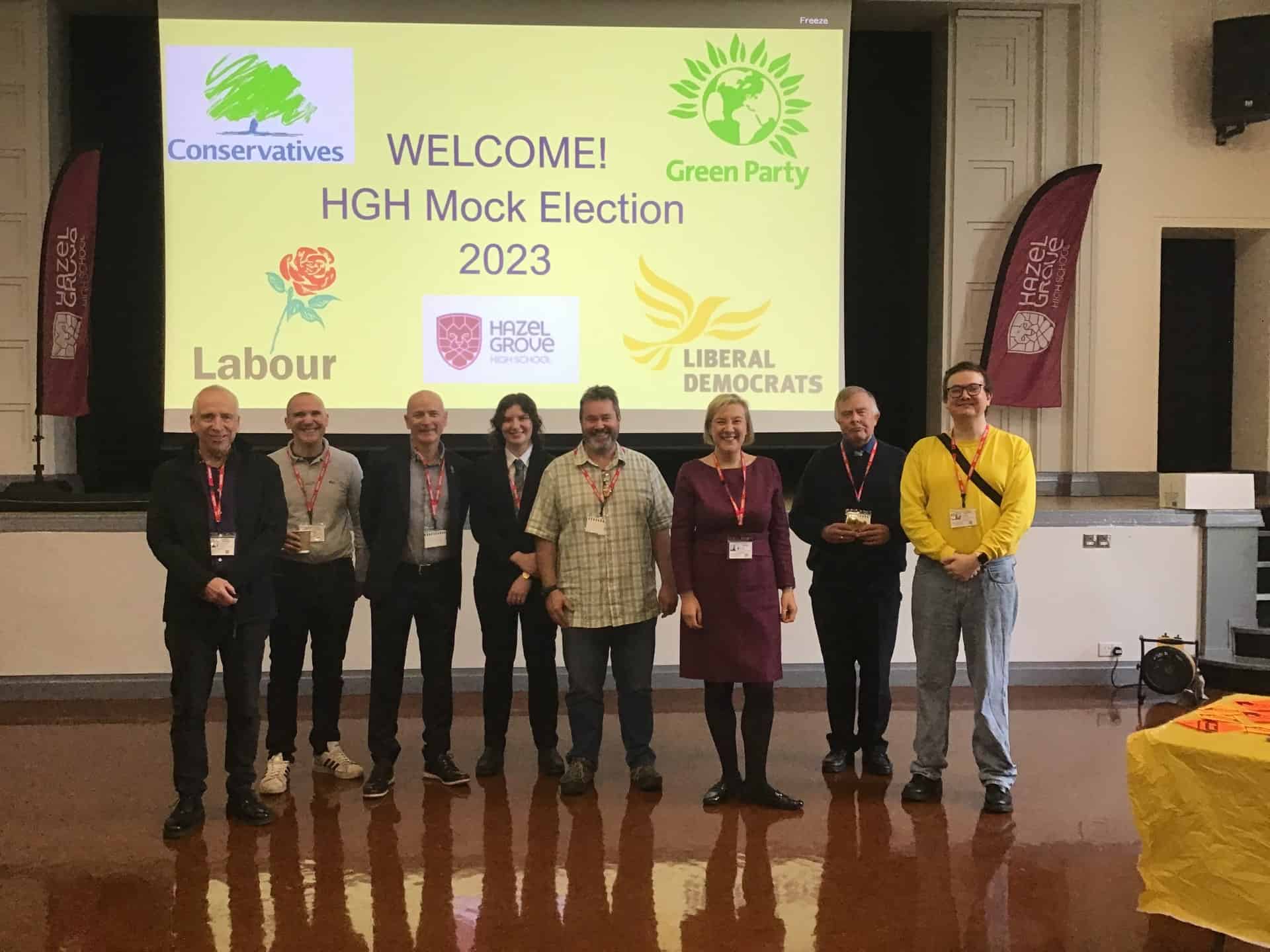 Local representatives from different political parties stand together at Hazel Grove High School in front of a screen which says: WELCOME! HGHS Mock Election 2023